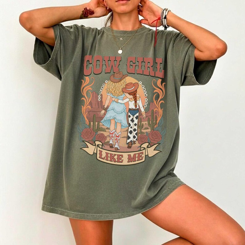 May include: An olive green short-sleeve t-shirt featuring a graphic print of two figures in cowboy hats walking through a desert landscape with cacti and roses. The text reads COW GIRL LIKE ME in a bold, arched font above and below the illustration.