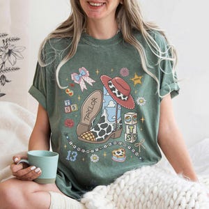 Op de afbeelding: Een saliegroen t-shirt met korte mouwen en een grafische print van een cowboylaars met koeienprint en de naam Taylor, een rode hoed, alfabetblokken, een strik en het getal 13, versierd met kleine sterretjes en bloemen.