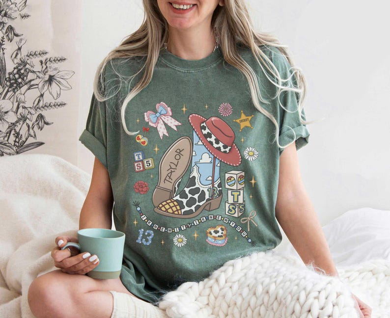 Op de afbeelding: Een saliegroen t-shirt met korte mouwen en een grafische print van een cowboylaars met koeienprint en de naam Taylor, een rode hoed, alfabetblokken, een strik en het getal 13, versierd met kleine sterretjes en bloemen.
