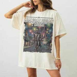 Op de afbeelding: Een gebroken wit t-shirt met korte mouwen en een grafische print van The Mandalorian en Grogu die door een straat lopen met een kasteel op de achtergrond onder kleurrijk vuurwerk. De tekst THE MANDALORIAN & GROGU staat boven de illustratie.