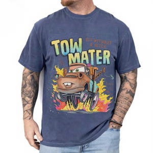 May include: A blue short-sleeve t-shirt featuring a graphic of the character Tow Mater from the movie Cars. The design includes the text "TOW MATER" and "OFF WITHOUT A HITCH" above the character, who is surrounded by orange and yellow flames.
