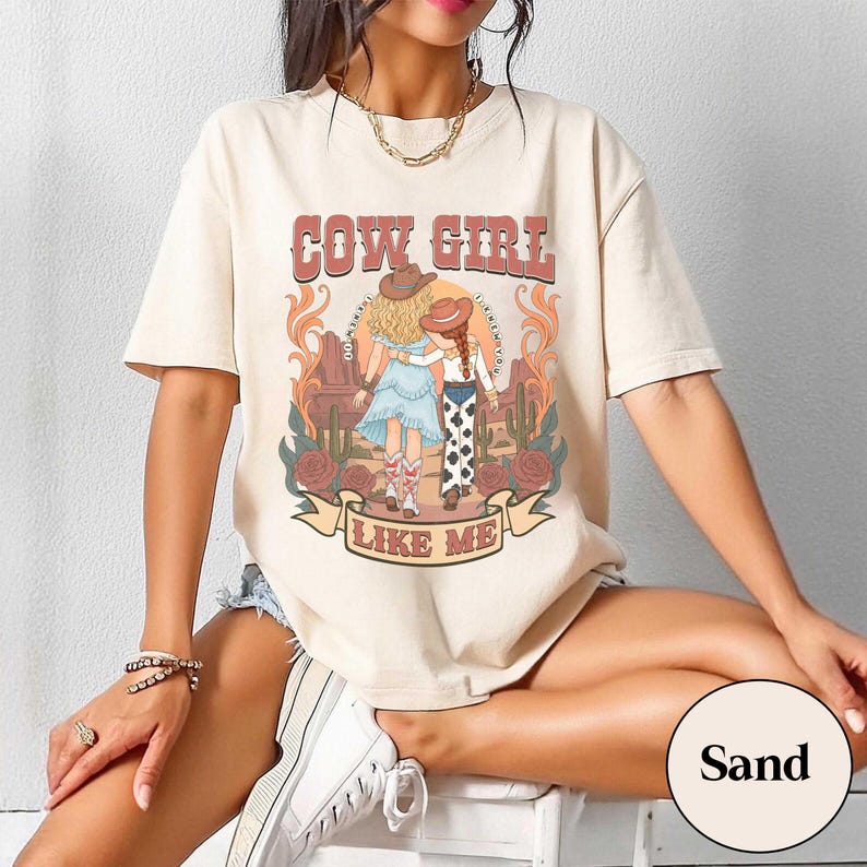 May include: A sand-colored short-sleeve t-shirt featuring a graphic print of two figures in cowboy hats walking through a desert landscape with cacti and roses. The text COW GIRL LIKE ME is printed in a bold, retro-style font above and below the illustration.