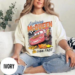 May include: Ivory-colored t-shirt featuring a graphic of Lightning McQueen from the movie Cars. The design includes the text "Lightning McQueen", "95", and "Think Fast". The shirt has a vintage, distressed look.
