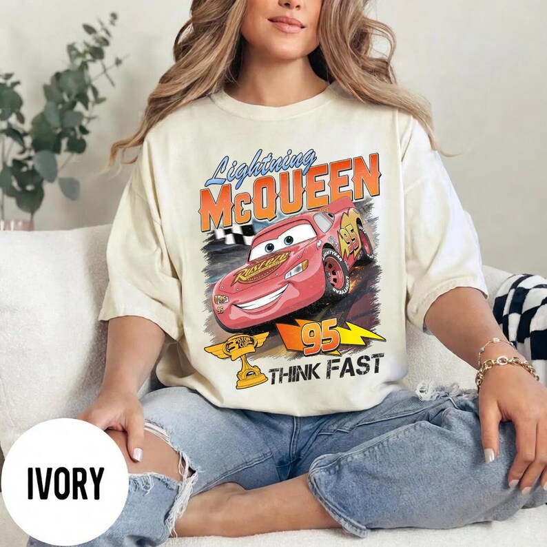 May include: Ivory-colored t-shirt featuring a graphic of Lightning McQueen from the movie Cars. The design includes the text "Lightning McQueen", "95", and "Think Fast". The shirt has a vintage, distressed look.