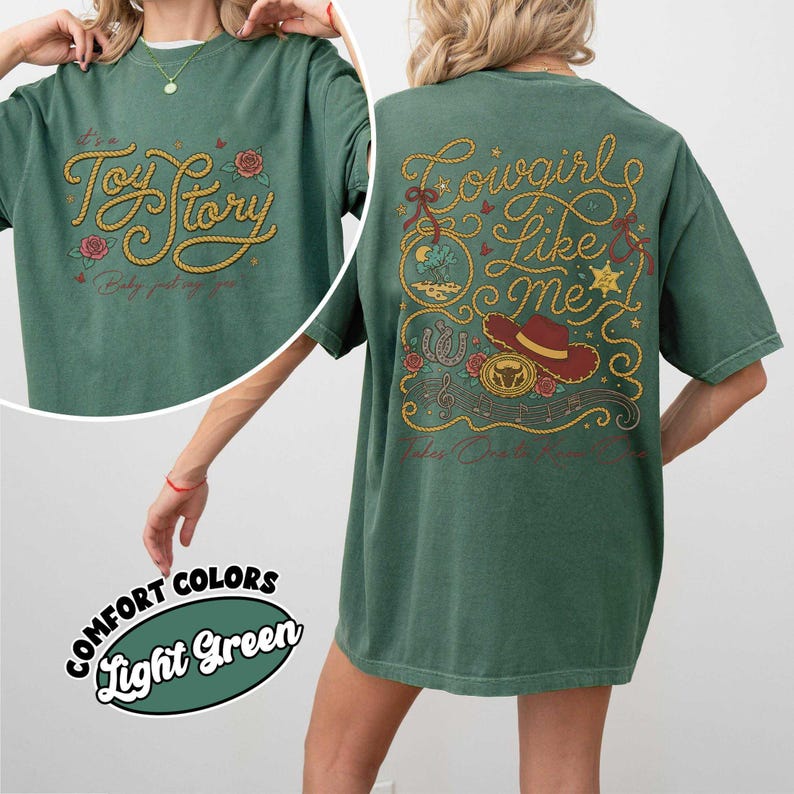 Puede incluir: Una camiseta de manga corta de color verde claro con un gr&aacute;fico en la espalda que incluye el texto "Cowgirl Like Me" en letras amarillas estilo cuerda, un sombrero de vaquero rojo, una herradura, rosas y notas musicales. El frente dice "It's a Toy Story".