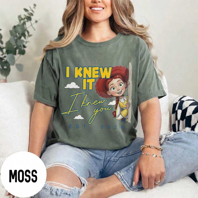 May include: A moss green short-sleeve t-shirt featuring a graphic of a cartoon cowgirl character with red hair and a hat. The text reads I KNEW IT in yellow block letters and I knew you in yellow script, with the text EST 2026 in blue below.