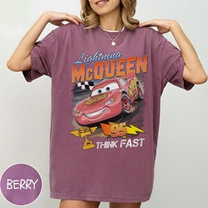 May include: Berry-colored t-shirt featuring a graphic of Lightning McQueen from the movie Cars. The design includes the text "Lightning McQueen", "95", and "Think Fast". The shirt has short sleeves and a crew neck.