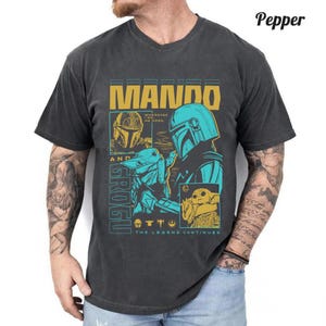 May include: A charcoal gray short-sleeve t-shirt featuring a graphic print of Mando and Grogu. The design includes the text MANDO, GROGU, WHEREVER I GO, HE GOES, and THE LEGEND CONTINUES, with yellow and teal illustrations of the characters.
