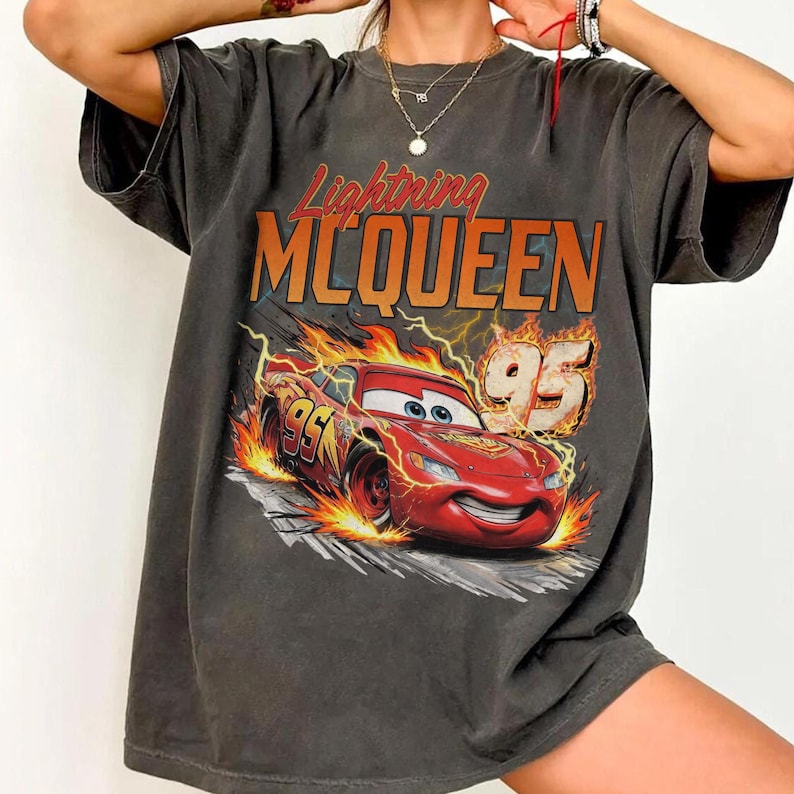 May include: A charcoal gray short-sleeve t-shirt featuring a graphic print of the red race car Lightning McQueen surrounded by orange and yellow flames. The text Lightning MCQUEEN and the number 95 are printed in bold orange and white lettering.