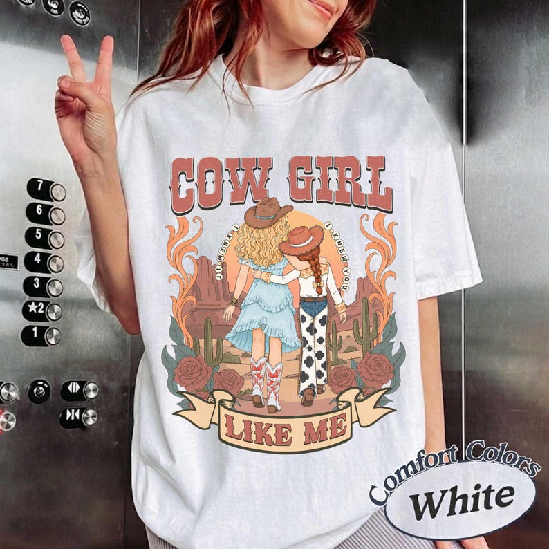 May include: A white t-shirt featuring a graphic print of two figures in cowboy hats standing in a desert landscape with cacti and roses. The text COW GIRL is printed above and LIKE ME is printed below. The figures wear western-style clothing.