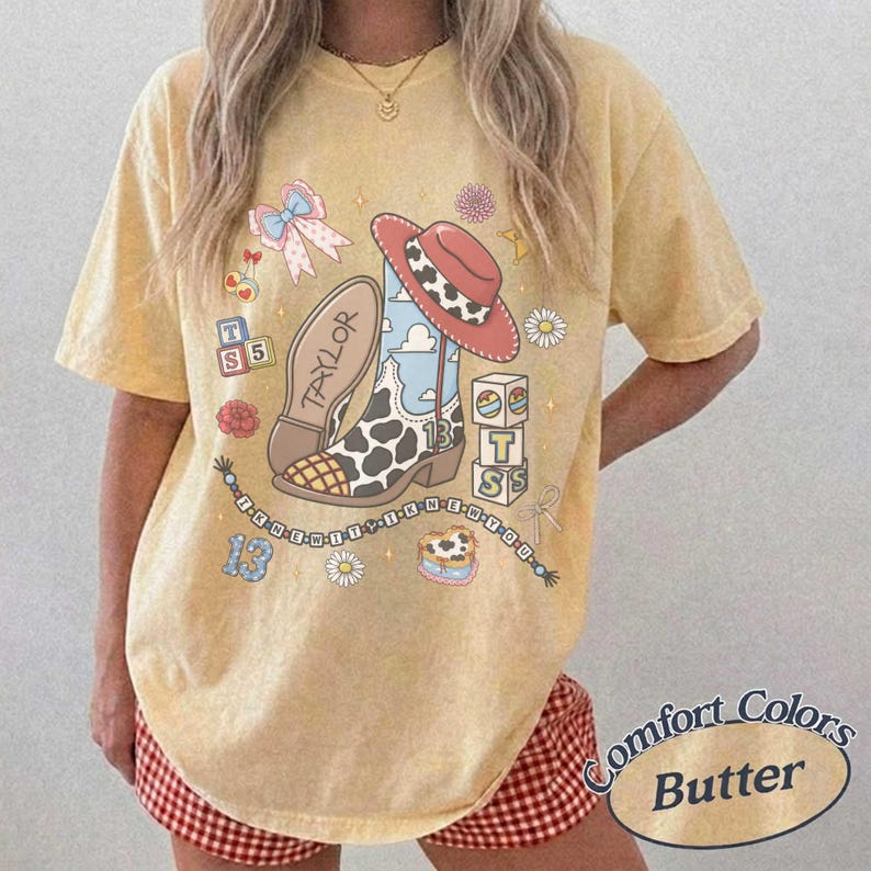 Op de afbeelding: Een geel t-shirt met korte mouwen en een grafische print van een cowboylaars met koeien- en wolkenpatroon, een rode hoed, alfabetblokken met de letters TS, een strik, bloemen en de tekst TAYLOR en I KNEW IT I KNEW YOU.