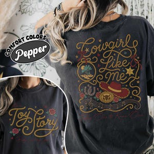 Puede incluir: Una camiseta gris carb&oacute;n con un gr&aacute;fico en la espalda que incluye letras de cuerda amarilla que dicen Cowgirl Like Me, un sombrero de vaquero rojo, una herradura, rosas y una estrella de sheriff. El frente muestra el texto Toy Story en cuerda.