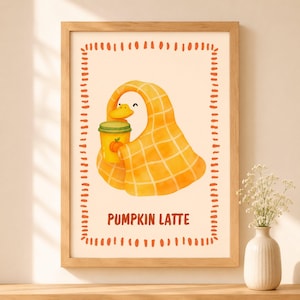May include: A framed wall art print featuring a white duck wrapped in a yellow plaid blanket, holding a yellow cup with a pumpkin icon. The text PUMPKIN LATTE is printed below the duck, all enclosed within a decorative orange dashed border.