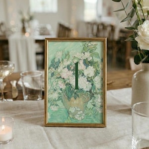 May include: A framed table number featuring a floral painting with a gold frame. The painting depicts white flowers in a vase with the number "1" in green. The table setting includes a candle, glasses, and a vase of white roses.