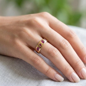 May include: A gold ring featuring oval red gemstones set across the band. The ring is on a finger, with a neutral background.
