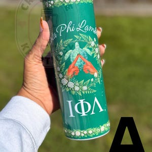 May include: A green tumbler with white floral accents and the text "Iota Phi Lambda." The design includes a turtle and hands forming a triangle. The tumbler is held in a hand.