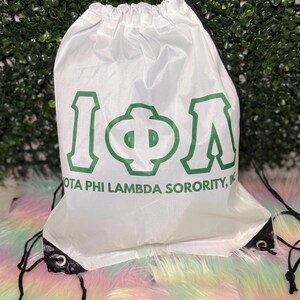 May include: White drawstring bag with green Greek letters and the text "IOTA PHI LAMBDA SORORITY, INC." Black drawstrings and eyelets. The bag is on a colorful, fluffy surface.
