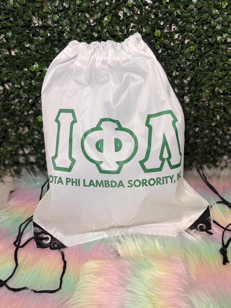 May include: White drawstring bag with green Greek letters and the text "IOTA PHI LAMBDA SORORITY, INC." Black drawstrings and eyelets. The bag is on a colorful, fluffy surface.