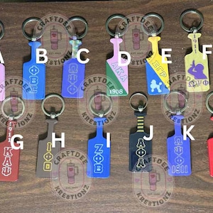 May include: A collection of keychains in various colors, each featuring a unique design and Greek letters. The keychains are attached to silver key rings. The keychains are made of acrylic or similar material.
