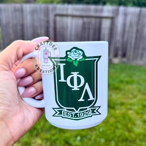 May include: A white ceramic mug featuring a dark green shield emblem with the Greek letters Iota Phi Lambda in white. Above the shield is a white rose, and below is a banner with the text EST. 1929. A circular logo with the text CRAFTDEE KRESTIONZ is visible.