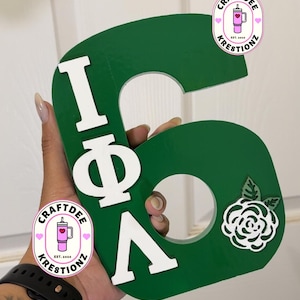 May include: A large, green wooden letter "G" with white Greek letters and a white rose design. The letter is held up in front of a white background. The logo "Craftdee Kreationz" is visible.