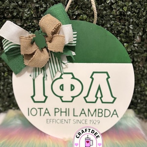 Puede incluir: Un letrero redondo de madera de dos tonos, con la parte superior verde y la inferior blanca. El letrero presenta las letras griegas Iota, Phi y Lambda en verde, con el texto "IOTA PHI LAMBDA EFFICIENT SINCE 1929". Un lazo con cintas verdes, blancas y de arpillera y un asa de cuerda completan el diseño.