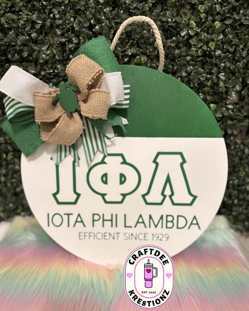 May include: A circular wooden sign, half green and half white, with the Greek letters Iota, Phi, and Lambda in green. The text reads "IOTA PHI LAMBDA EFFICIENT SINCE 1929." A bow made of green, white, and burlap ribbons and a rope handle are attached to the sign.