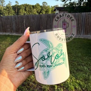 May include: White Soror Iota Phi Lambda mug with a green sea turtle design. The mug has a handle and a lid. The text "Soror" is written in a script font, and the words "Iota Phi Lambda" are below the turtle.