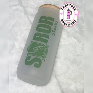 May include: A frosted glass tumbler with a bamboo lid. The tumbler features green text that reads "SOROR" and "Tanitra", along with a crest and "EST. 1929". A logo for "Craftdee Kreztionz" is visible in the upper right corner.