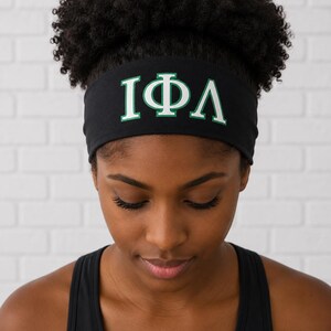 May include: A black fabric headband featuring the Greek letters Iota, Phi, and Lambda printed in white with a green outline. The headband is wide and designed to fit securely around the head, displaying the letters clearly across the front center.