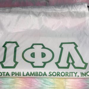 May include: White drawstring bag with the Greek letters Iota, Phi, and Lambda in green, along with the text "IOTA PHI LAMBDA SORORITY, INC." The bag has black drawstrings and is set against a colorful, fluffy background.