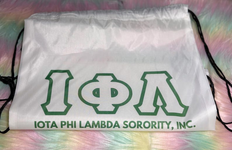 May include: White drawstring bag with the Greek letters Iota, Phi, and Lambda in green, along with the text "IOTA PHI LAMBDA SORORITY, INC." The bag has black drawstrings and is set against a colorful, fluffy background.