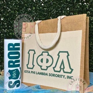 May include: A tan burlap tote bag with white handles and a cream-colored front panel featuring the dark green Greek letters Iota Phi Lambda and the text IOTA PHI LAMBDA SORORITY, INC. Next to it is a white cylindrical tumbler with matching green text.
