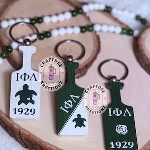 May include: Three wooden keychains in white and green, shaped like paddles. Each keychain features Greek letters, a turtle or a rose, and the year 1929. The keychains are displayed on a wooden slice with a beaded garland.