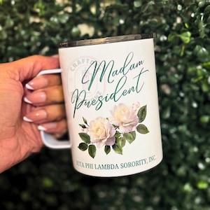 May include: A white mug featuring the dark green cursive text Madam President above a cluster of pale pink roses with green leaves. The base of the mug displays the text IOTA PHI LAMBDA SORORITY, INC. in dark green capital letters.