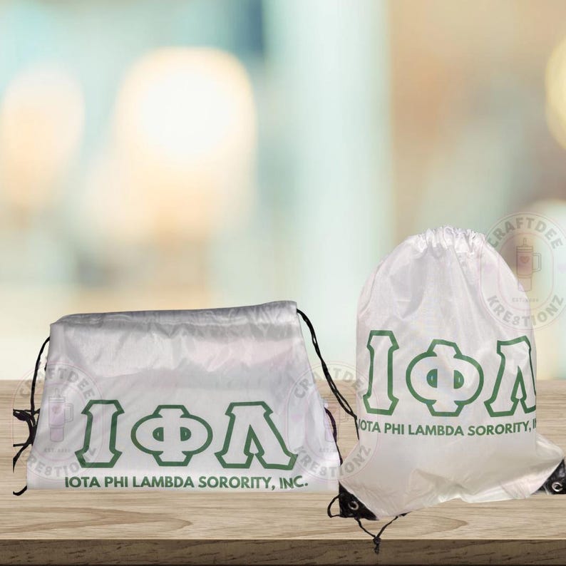 May include: Two white drawstring bags with the Greek letters I&Phi;&Lambda; in green. The text "IOTA PHI LAMBDA SORORITY, INC." is printed below the letters. The bags are on a wooden surface.