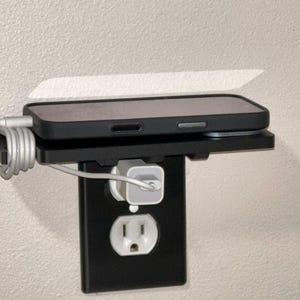 Puede incluir: Estante negro para toma de corriente con un teléfono inteligente encima. Un cable de carga blanco está enchufado al teléfono y enrollado. El estante está montado en una cubierta de toma de corriente negra.