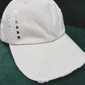 May include: A light beige baseball cap with a distressed brim. The cap features a series of heart-shaped designs in various shades of brown and white on the side. The cap is made of a soft fabric.