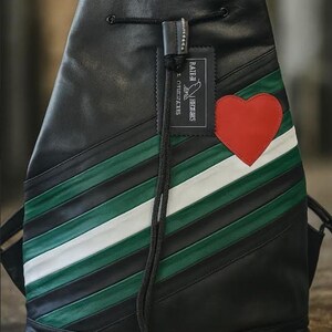 May include: Black leather backpack with a diagonal stripe design in green and white. A red heart is stitched on the front. The backpack has a drawstring closure and a tag that reads "RATHI HERMES".