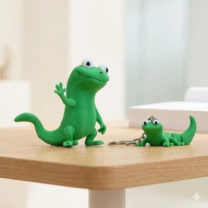 May include: Two green lizard figurines. One is a large, upright lizard with its arm raised in a wave. The other is a smaller lizard keychain. Both have large, round eyes and are on a wooden surface.