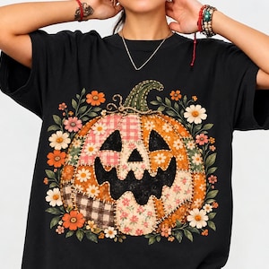 May include: A black short-sleeved t-shirt featuring a large central graphic of a jack-o'-lantern pumpkin. The pumpkin is designed with various patchwork patterns including plaid, gingham, and floral prints, surrounded by orange and white flowers.