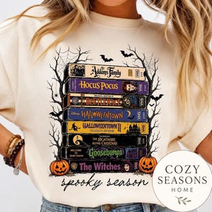 May include: A cream-colored t-shirt featuring a graphic of stacked VHS tapes labeled with movie titles including The Addams Family, Hocus Pocus, Beetlejuice, Halloweentown, The Nightmare Before Christmas, Goosebumps, and The Witches, framed by trees and pumpkins.