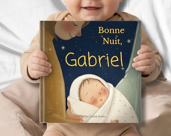 May include: A smiling baby holds a children's book titled "Bonne Nuit, Gabriel." The book cover features a nighttime scene with a sleeping baby and the text in yellow. The book is from Blue Trunk Books.