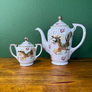 May include: A white porcelain teapot and a matching sugar bowl, both featuring a gold and black dragon motif with red accents. The teapot has a lid with a gold finial, a curved handle, and a spout. The sugar bowl has two side handles and a lid.
