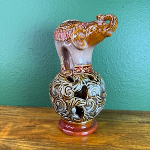 May include: A ceramic elephant figurine with a glossy brown finish and a raised trunk. The base is a spherical, hollowed-out structure featuring intricate, swirling relief patterns in shades of brown, cream, and orange with cut-out openings.