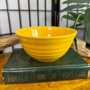 May include: A bright yellow ceramic bowl with a ribbed exterior. The bowl is sitting on top of a dark green book with gold lettering and decorative elements. The background includes a wooden element and green foliage.
