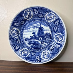 May include: A round ceramic plate featuring a blue and white transferware pattern. The center depicts a Dutch windmill landscape with buildings and figures. The wide border is decorated with intricate floral motifs and scrolling foliage designs.