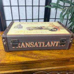May include: A brown decorative box with a vintage travel theme. The top features an image of a car and the text "WEEK END &agrave; Londres". The front of the box reads "TRANSATLANT". The box has metal clasps and brown leather trim.