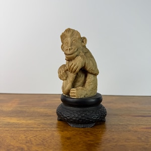 May include: A beige, carved monkey figurine in a seated position, holding a small object in its hands. The monkey has detailed fur texture and a smiling expression. It is mounted on a circular, dark brown, textured wooden display base.