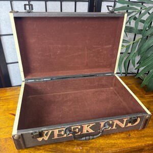 May include: An open, vintage-style suitcase with the word "WEEKEND" printed on the front. The suitcase is brown with a cream-colored trim and a dark brown interior. It has a metal handle and clasp.